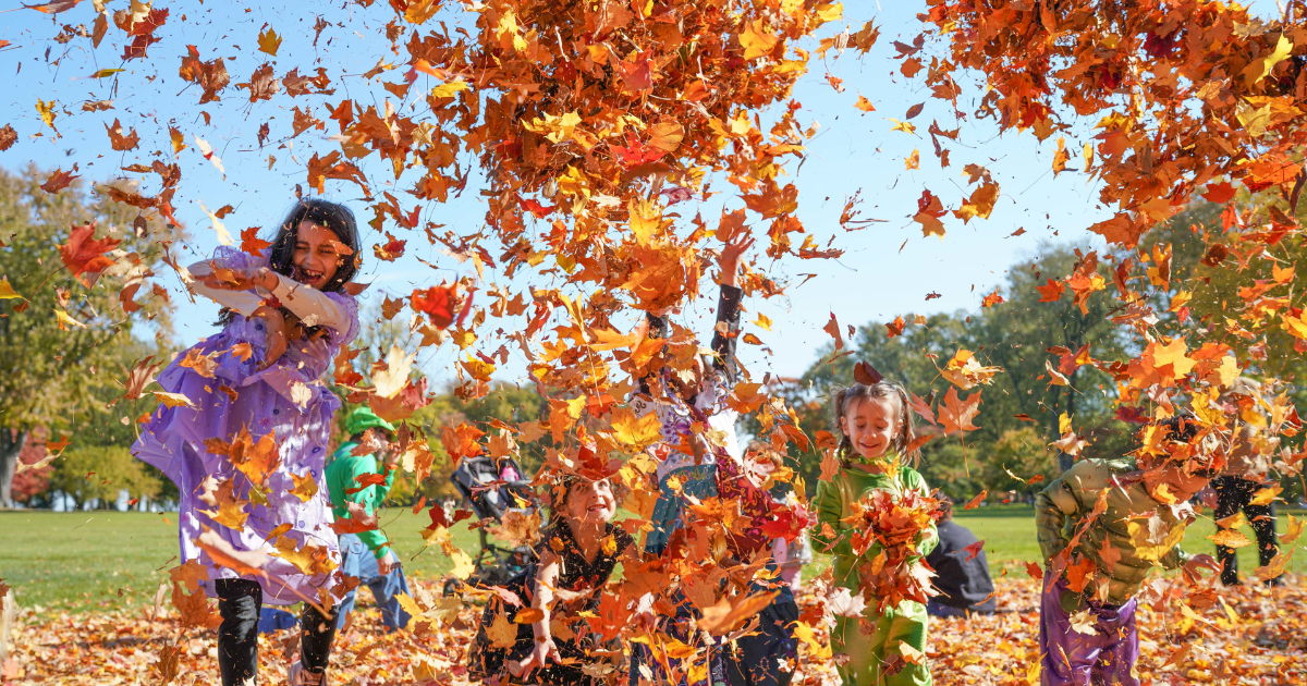 Children play in a pile of autumn leaves, tossing them into the air at Ford House’s Little Goblins event.