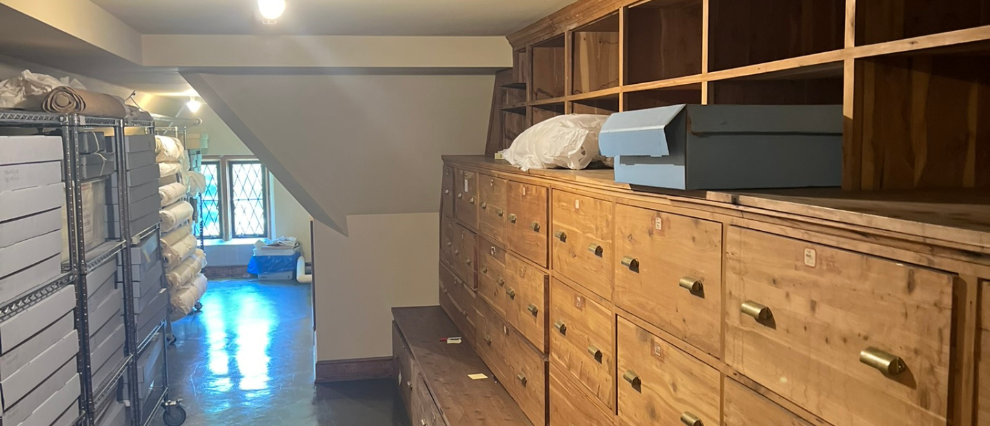 A well-organized storage room with rows of labeled wooden drawers on the right and metal shelving units holding archival boxes and rolled textiles on the left. Light from a window at the end of the room illuminates the polished floor.