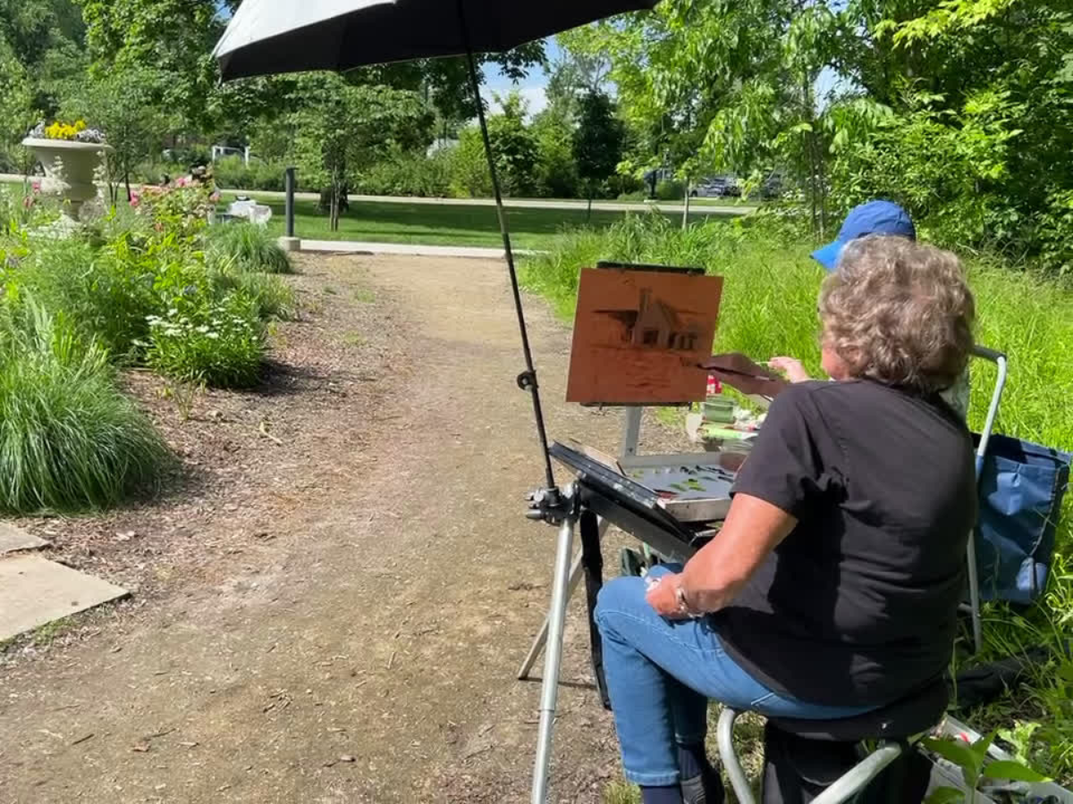 Artist painting outdoors during Ford House Paint Out, seated under an umbrella with an easel set up along a garden path.