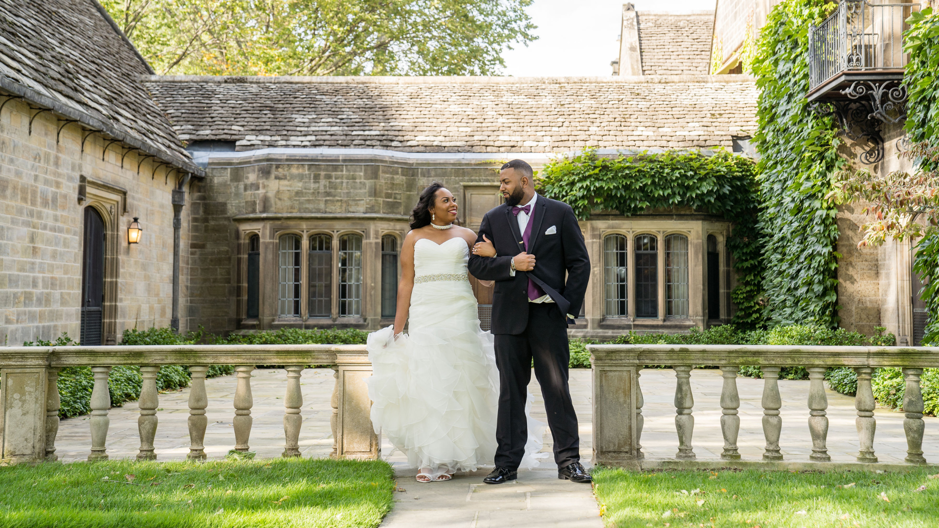 A newlywed couple walks arm in arm in a beautiful historic stone courtyard with ivy-covered walls and elegant windows, smiling at each other on their wedding day.