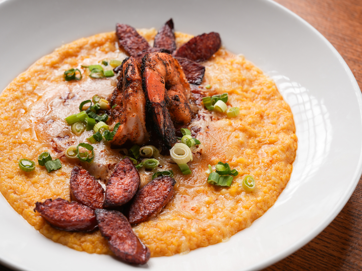 A bowl of creamy cheddar grits topped with grilled shrimp, smoked sausage, scallions, and melted butter.