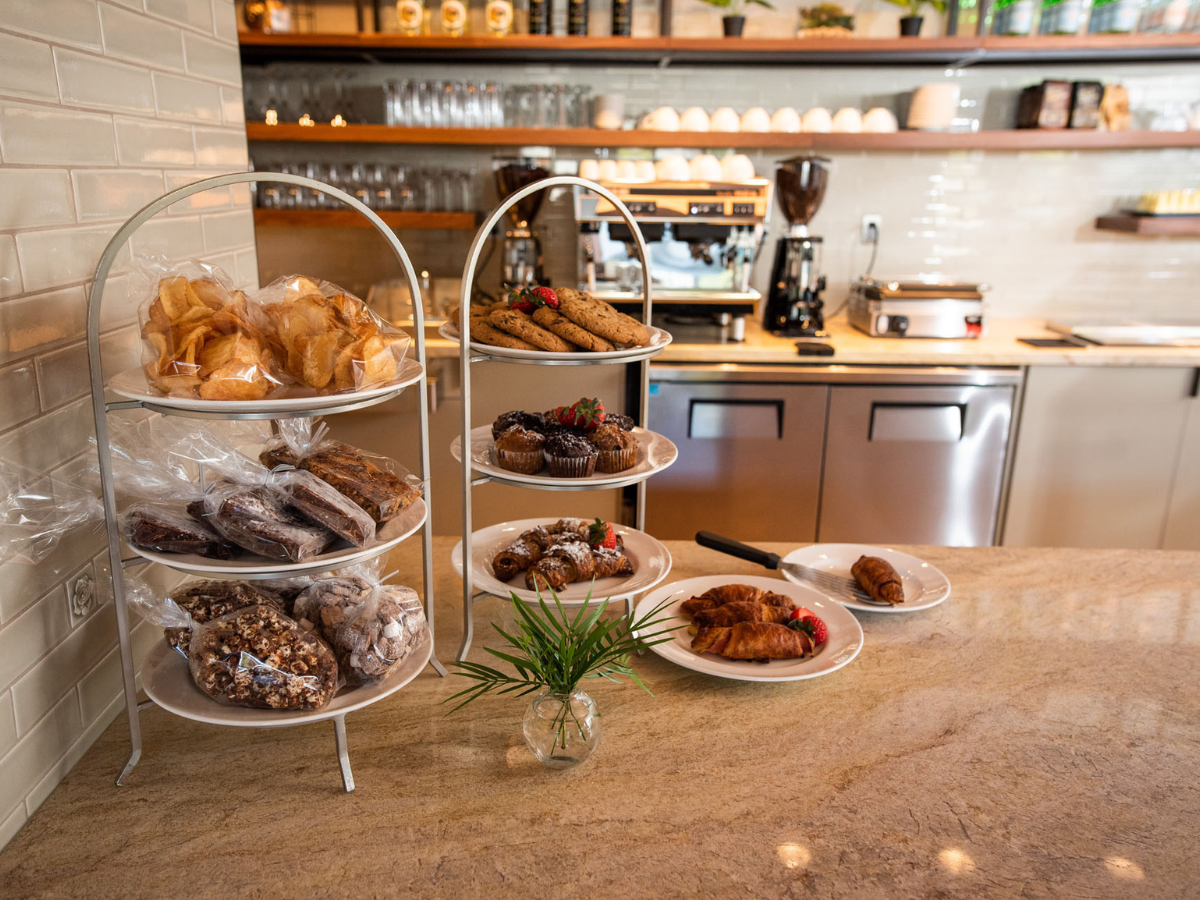 Display of pastries, cookies, brownies, and packaged snacks arranged on tiered stands at a café counter.