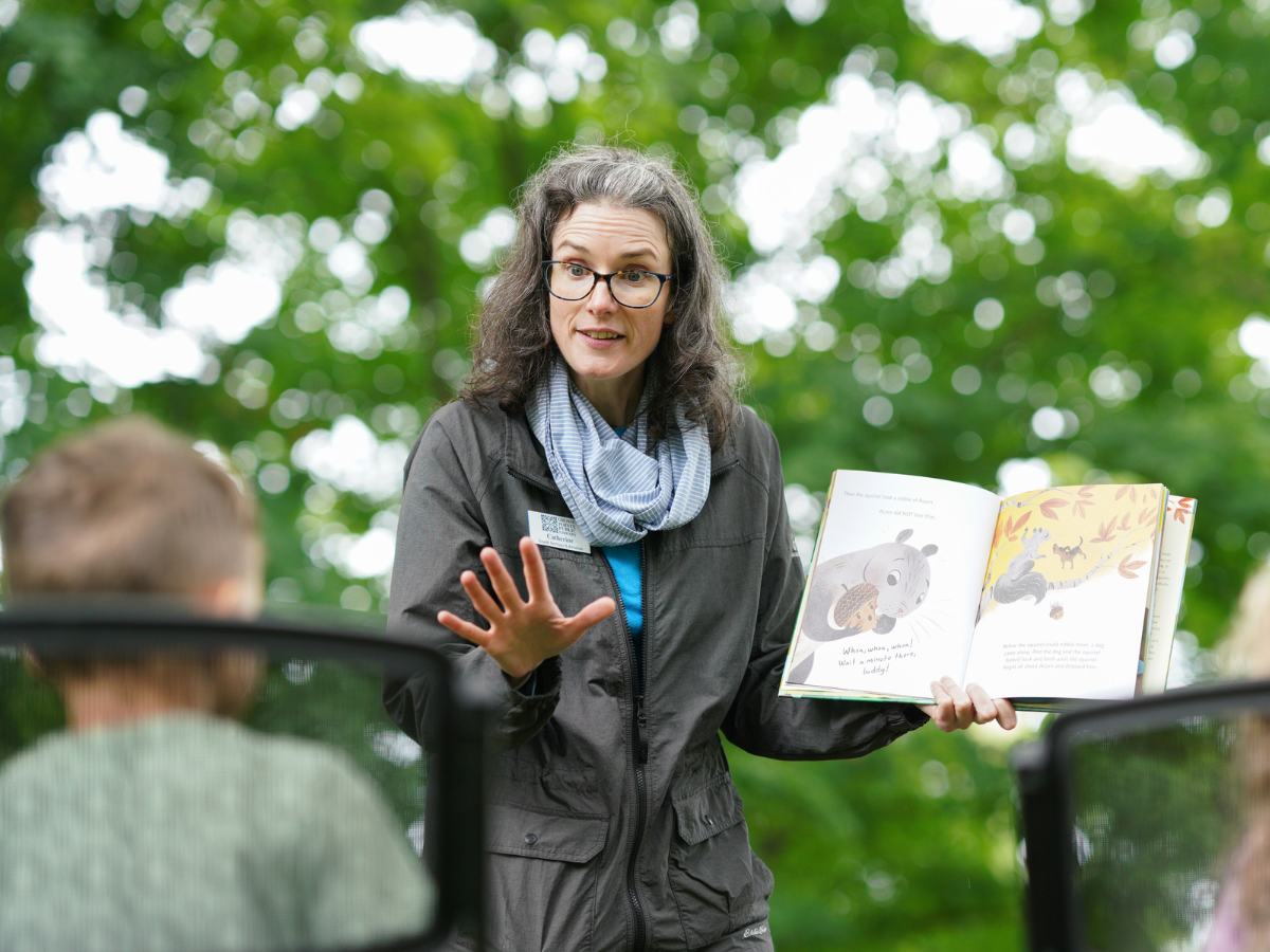 Educator holding open a picture book and speaking to children outdoors at Story Festival.