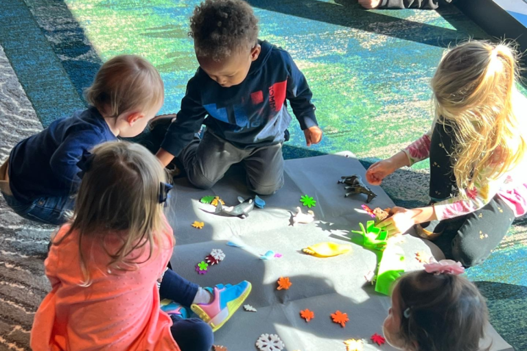 A diverse group of young children sits on a soft mat in a sunlit room, exploring colorful animal figures and nature-themed toys during Storytime at Ford House.