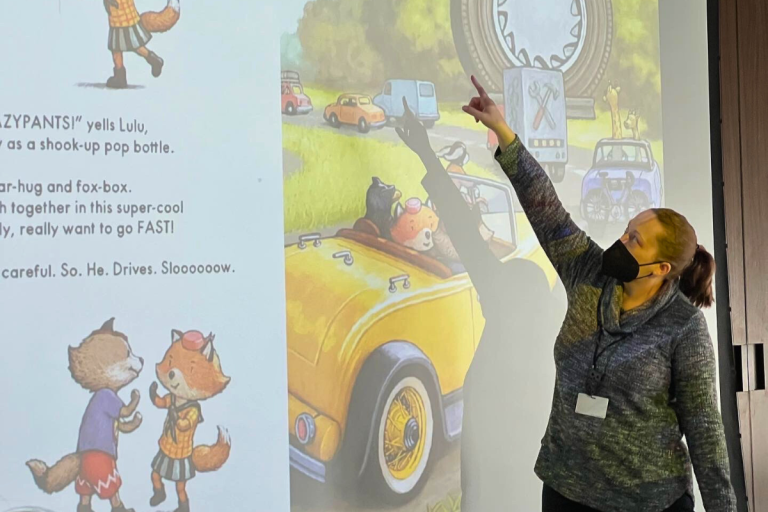A woman wearing a mask and cozy sweater gestures toward a large screen projection of a children’s picture book featuring cartoon animals in a yellow convertible.