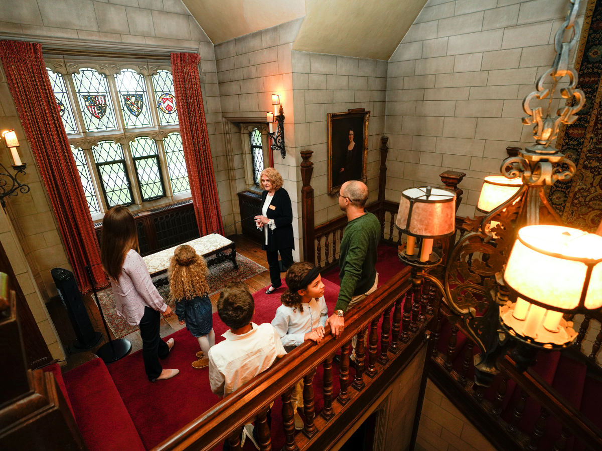 A Ford House tour guide leads a family through the historic Main Residence, sharing stories in a grand hallway with stained glass windows and elegant details.